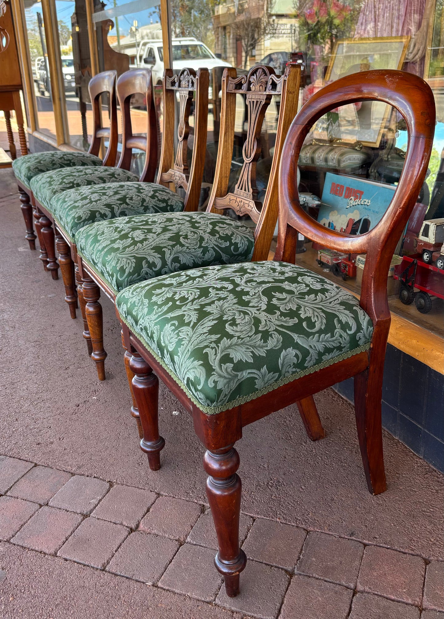 Row of wooden chairs with green patterned cushions on a brick pavement.