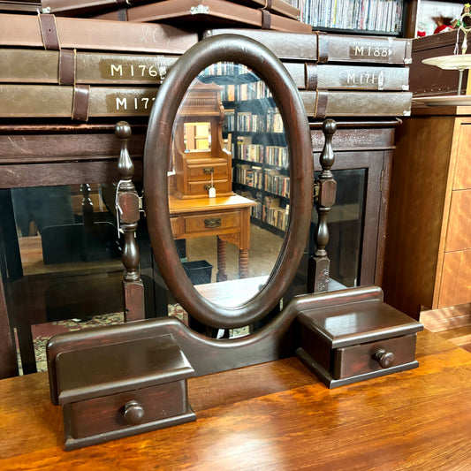 Wooden vanity mirror with drawer on a wooden surface, with a bookshelf in the background.