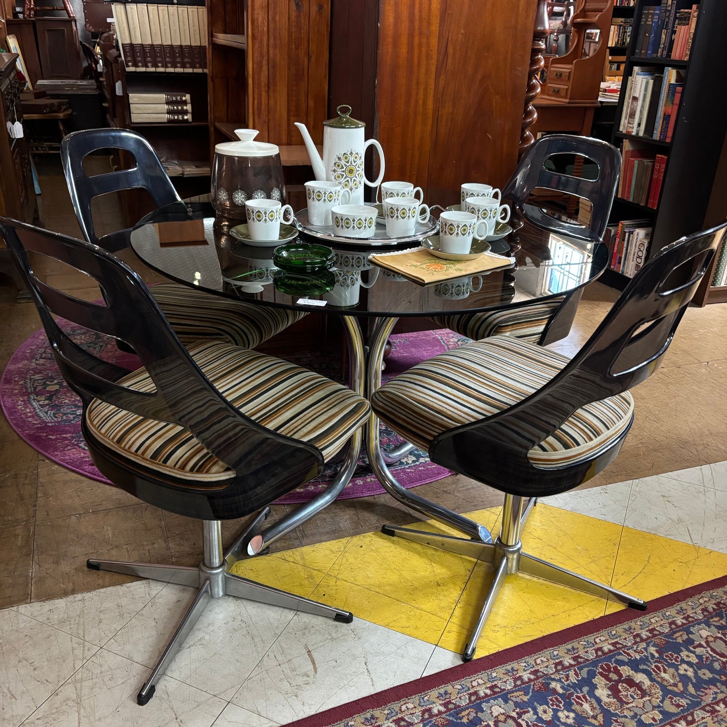 Round glass dining table with black chairs and striped cushions in a room with wooden furniture and books.