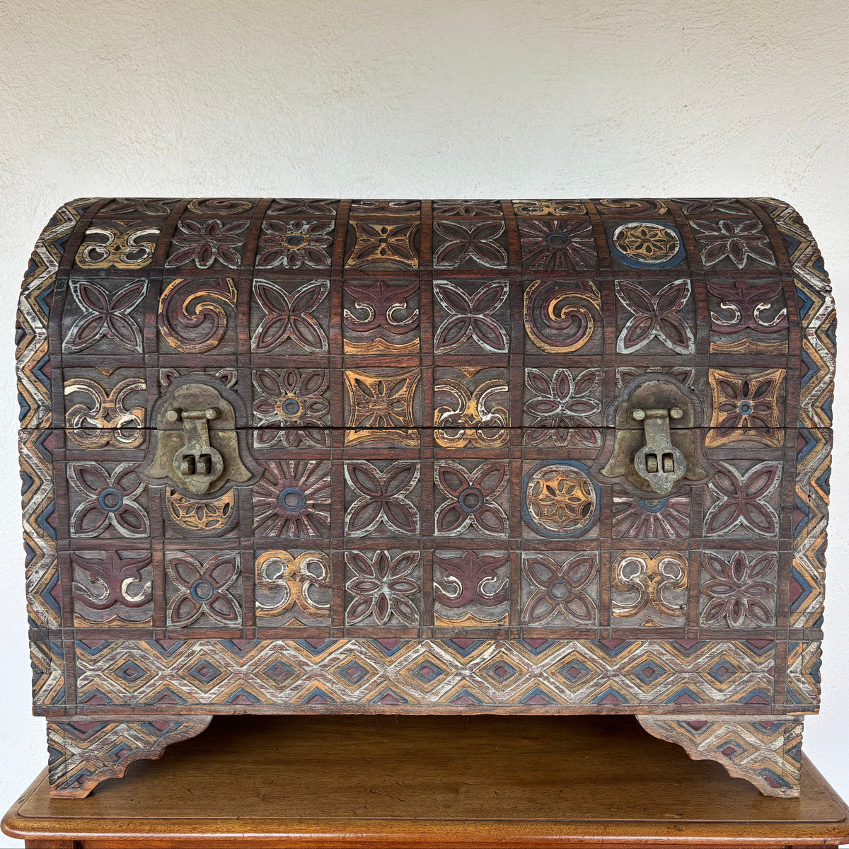 An antique carved wooden chest with intricate geometric and floral patterns, featuring two metal latch locks, displayed on a wooden table against a textured off-white wall.